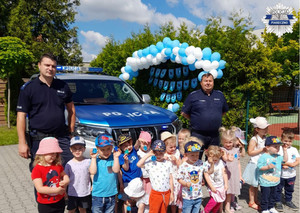 Zdjęcie grupki dzieci w scenerii pikniku rodzinnego - w tle wiązki baloników, obok dzieci stoi dwóch umundurowanych policjantów.