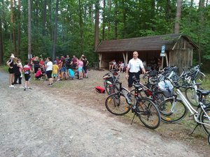 Zdjęcie leśnego kempingu, w tle grupa osób, na pierwszym planie stojące rowery i uśmiechnięty policjant umundurowany w biała koszulkę i krótkie spodenki.