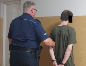 Zdjęcie stojących twarzami do ściany umundurowanego policjanta i mężczyzny z kajdankami na rękach.