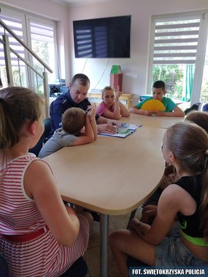 Zdjęcie sali, w której przy stoliku siedzi grupka dzieci, a wśród nich umundurowany policjant.