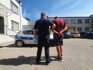 Dziedziniec komendy policji, na którym widoczni są odwróceni tyłem: umundurowany policjant i mężczyzna ze skutymi kajdankami rękoma.