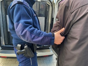 Zdjęcie przedstawiające środkową cześć sylwetki umundurowanego policjanta przytrzymującego za przedramię mężczyznę w kurtce.