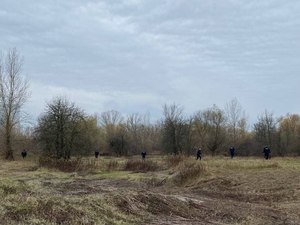 Zdjęcie zakrzaczonego terenu jesienią, w dali widoczny szpaler umundurowanych policjantów.