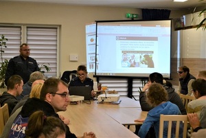 Sala konferencyjna, siedząca grupa osób, w tle dwójka umundurowanych policjantów.