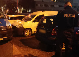 Parking nocą, na pierwszym planie odwrócony tyłem umundurowany policjant.