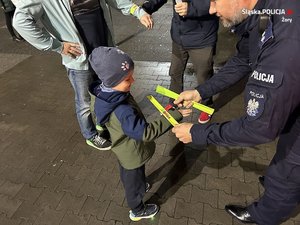 Kucający umundurowany policjant zakładający na rękę dziecka opaskę odblaskową.