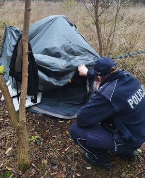 Prowizoryczny namiot z czarnej folii, przy którym kuca umundurowany policjant.