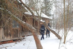 Drewniany budynek z zaroślami w zimowej scenerii, w oddali dwójka umundurowanych policjantów.