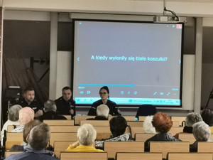 Sala konferencyjna z siedzącymi seniorami, za stołem trójka umundurowanych policjantów