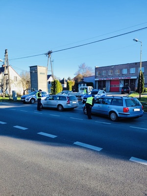 Ulica, na której zaparkowane są dwa radiowozy i dwa samochody obok nich dwóch umundurowanych policjantów.