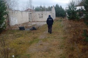 Opuszczony biały budynek bez dachu na tle zarośli oraz idący w jego stronę umundurowany policjant.