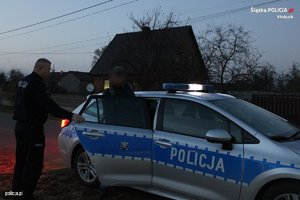 Wiejska droga, na której o zmroku stoi radiowóz, do którego wsiada niedbale ubrany mężczyzna, a obok stoi umundurowany policjant