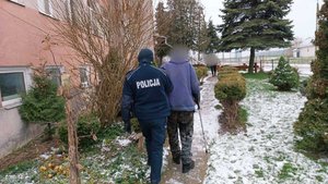 Mężczyzna oraz umundurowany policjant idący wzdłuż ściany budynku, na chodniku widoczny śnieg.