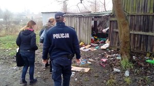 Ruiny drewnianych zabudowań przy których widoczne są dwie kobiety po cywilnemu i umundurowany policjant.