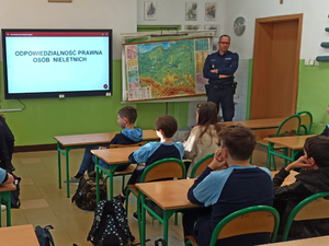 Sala szkolna wypełniona dziećmi w ławkach szkolnych. W tle umundurowany policjant prowadzący prelekcję.