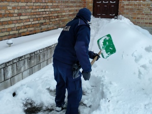 Umundurowany policjant odrzuca śnieg przed budynkiem.