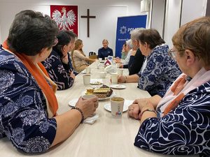 Sala konferencyjna wypełniona seniorami siedzącymi przy stole, na którym widoczna jest herbata i ciastka. U szczytu stołu umundurowana, uśmiechnięta policjantka.