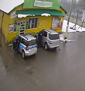 Zdjęcie z monitoringu pokazujące z góry wolnostojący sklep przed którym stoi zaparkowany radiowóz policyjne oraz auto osobowe w kolorze srebrnym.