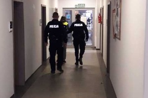 Dwaj umundurowani policjanci prowadzący między sobą korytarzem mężczyznę.