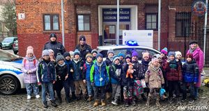 Grupa dzieci z opiekunką oraz dwójką umundurowanych policjantów na tle budynku komisariatu Policji z czerownej cegły.