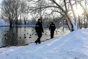 Dwaj umundurowani policjanci przy oczku wodnym w parku. Sceneria zimowa.