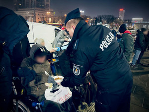 Plac Defilad w Warszawie w scenerii nocnej. Na pierwszym planie odwrócony tyłem umundurowany policjant podający jednorazową miseczkę z jedzeniem mężczyźnie na wózku inwalidzkim.