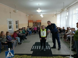 Sala przedszkolna wypełniona dziećmi na środku umundurowany policjant zakłada dziecku żółtą kamizelkę odblaskową.