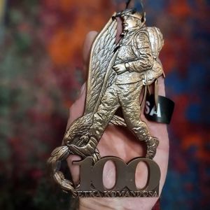 Metalowa leżąca na dłoni statuetka żołnierza na tle skrzydeł, w dolnej części napis &quot;Setka komandosa&quot; oraz liczba 100.