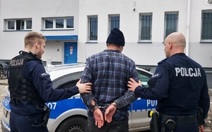 Mężczyzna przytrzymywany przez dwóch umundurowanych policjantów przed budynkiem komendy.