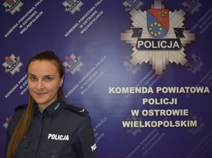 Umundurowana policjantka stojąca na tle granatowego banera Komendy Powiatowej Policji w Ostrowie Mazowieckim.