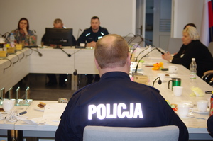 Ludzie siedzący przy stole konferencyjnym, na pierwszym planie odwrócony tyłem umundurowany policjant.