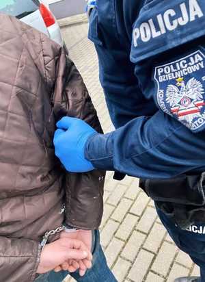Zbliżenie ręki umundurowanego policjanta przytrzymującego za rękę skutego kajdankami mężczyznę.