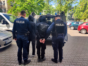 Dwóch widzianych z tyłu umundurowanych policjantów wprowadzających do więźniarki dwóch mężczyzn w czarnych bluzach dresowych.