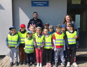 Grupka dzieci ubranych w żółte kamizelki odblaskowe na tle budynku komisariatu. W tle umundurowany policjant i ich opiekunka.