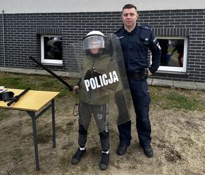 Umundurowany policjant oraz chłopiec w policyjnym kasku z przyłbicą i tarczą.