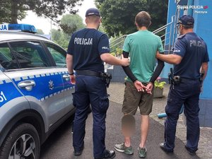 Odwróceni tyłem dwaj policjanci i mężczyzna ze skutymi z tyłu rękami obok budynku komisariatu i radiowozu.