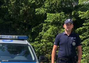 Umundurowany policjant przy radiowozie na tle szpaleru świerków