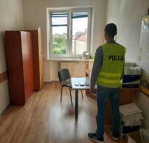 Policjant ubrany w żółtą kamizelkę odblaskową stojący tyłem w pomieszczeniu biurowym.