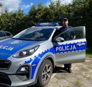 Policjant w otwartych drzwiach radiowozu na tle zieleni.