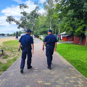 Dwóch umundurowanych policjantów idących alejką na tle zieleni widzianych tyłem