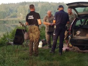 Brzeg jeziora gdzie stoją obok samochodu terenowego trzej mężczyźni w mundurach.