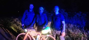 Zdjęcie w scenerii nocnej, trzy sylwetki stojące obok roweru.