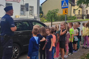 Grupka dzieci na chodniku obok samochodu, obok dzieci policjant