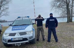 Dwaj mężczyźni, w tym jeden umundurowany policjant obok radiowozu stojącego nad brzegiem zbiornika wodnego