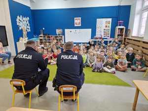 Sala z siedzącą na wykładzinie grupką dziećmi i dwoma umundurowanymi policjantami