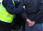 Zbliżenie pleców mężczyzny przytrzymywanego przez policjanta, widoczne skute kajdankami ręce