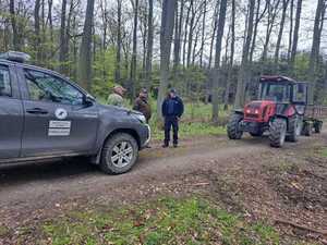 Umundurowany Policjant i dwóch strażników leśnych,  na pieszym planie samochód osobowy w oddali traktor rolniczy.
