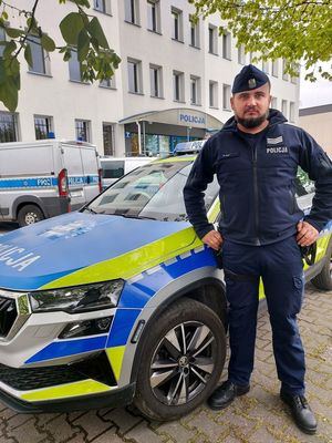 Umundurowany policjant z brodą obok radiowozu na tle budynku policji i zieleni