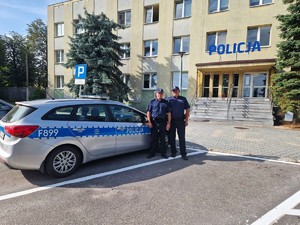 Dwoch umundurowanych policjantów obok radiowozu na tle budynku Policji