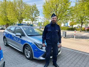 Uśmiechnięty, umundurowany policjant obok radiowozu, na tle zieleni miejskiej